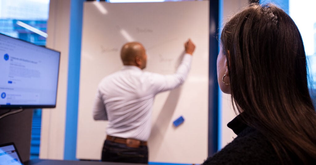 foto man en vrouw in kantoor. De vrouw zit achter de computer en kijkt naar de man. De man staat te schrijven op een whiteboard.