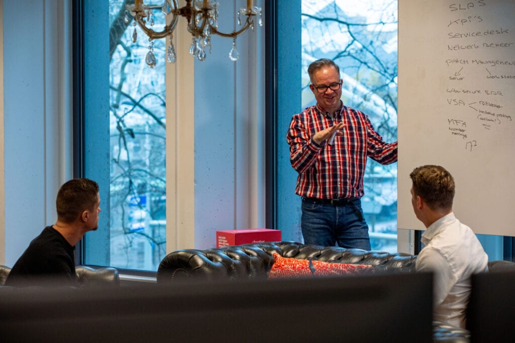 De afbeelding laat zien 3 mannen in comfortabel overleg op kantoor. Één staan bij het whiteboard de andere twee zitten op een zwarte stoel.