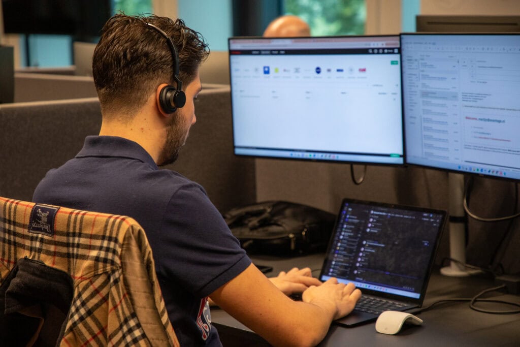 Man aan het werk achter drie computer schermen met headset op.