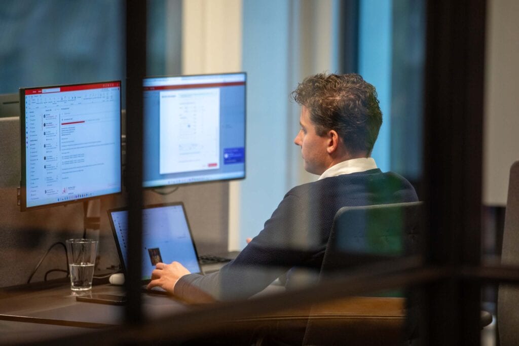Man werkt aan het bureau op het kantoor. Zichtbaar zijn drie werk schermen.