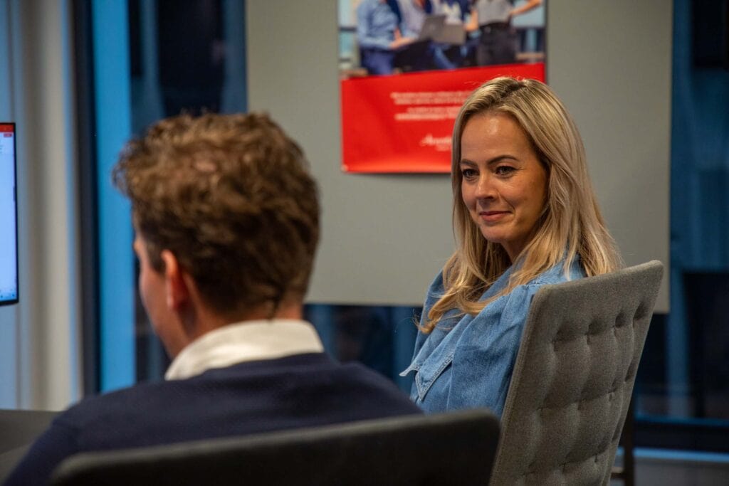 Foto van man en vrouw op kantoor. De vrouw kijkt naar de man. Ze zitten op stoelen.