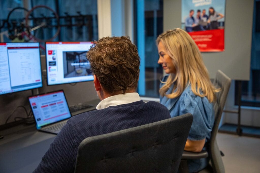 Foto van twee collega's op het kantoor. Ze zitten aan een bureau en kijken naar drie schermen.
