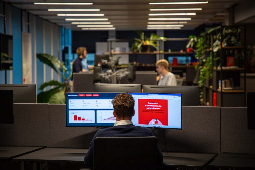 Man aan het werk in het kantoor. Hij zit achter twee beeldschermen. In de achtergrond zijn collega's aan het werk.