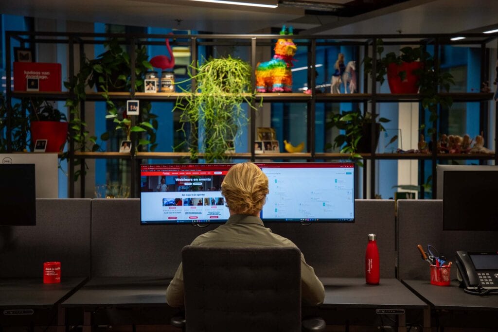 Foto van man op kantoor. Hij zit aan het bureau achter twee schermen met zichtbaar de Avantage website pagina Webinars en events & zijn agenda. Op de achtergrond is een kast met vele verschillende spullen te zien.