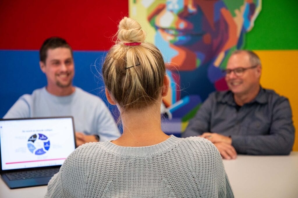 Vrouw zit met laptop tegenover twee mannen in de achtergrond.