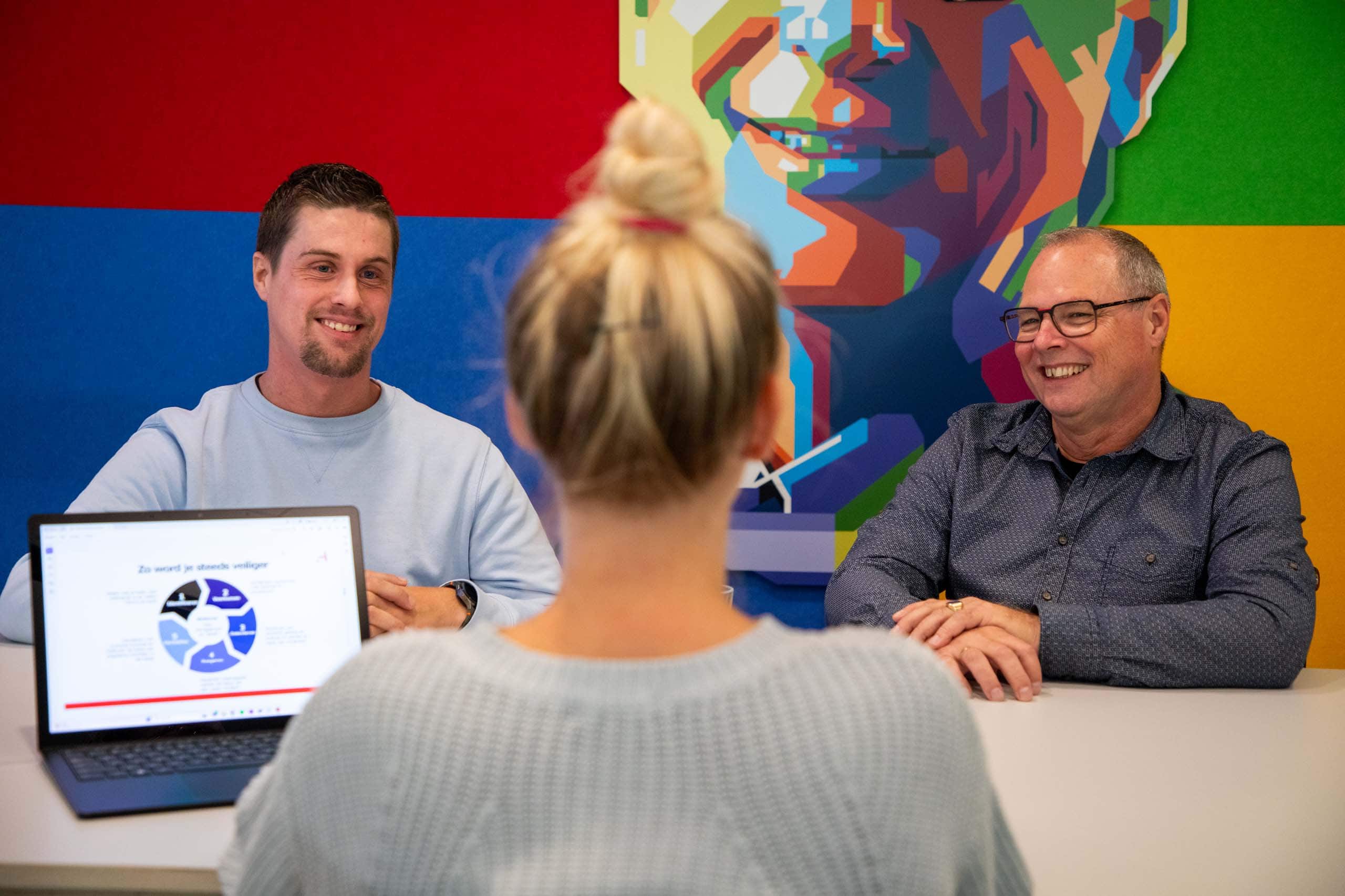Werkoverleg op kantoor met drie mensen. De twee mannnen kijken naar de vrouw, De vrouw heeft een laptop voor zich.