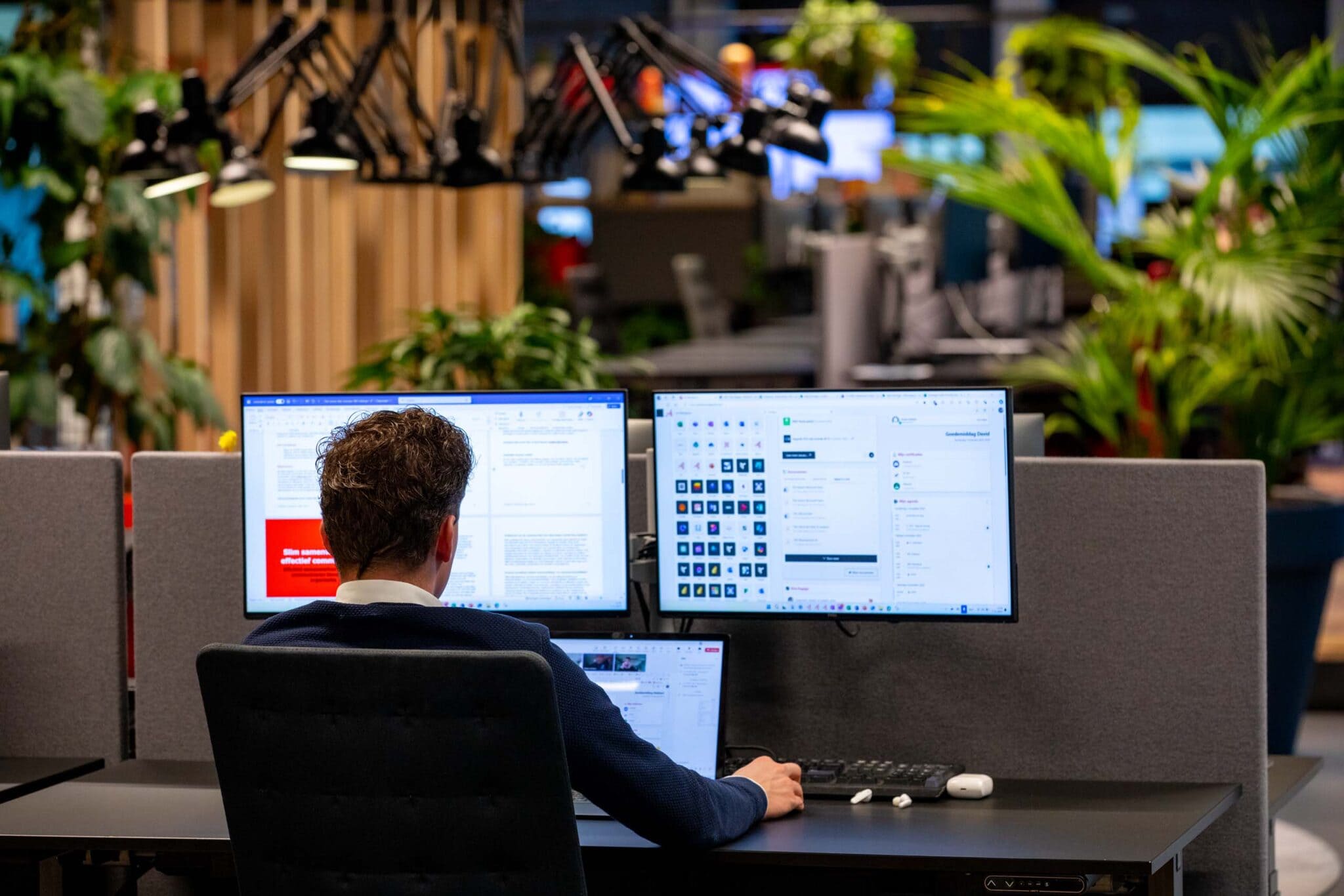 Foto van man op kantoor werkt achter drie schermen. Op de achtergrond zijn verschillende planten te zien.