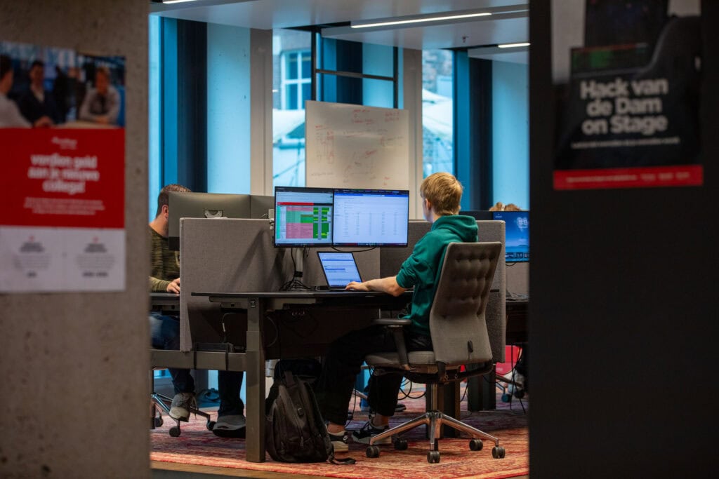 Man op kantoor aan het werk achter een bureau. Te zien zijn twee vervaagde poster op muren en op het bureau staat een laptop met twee extra beeldschermen.
