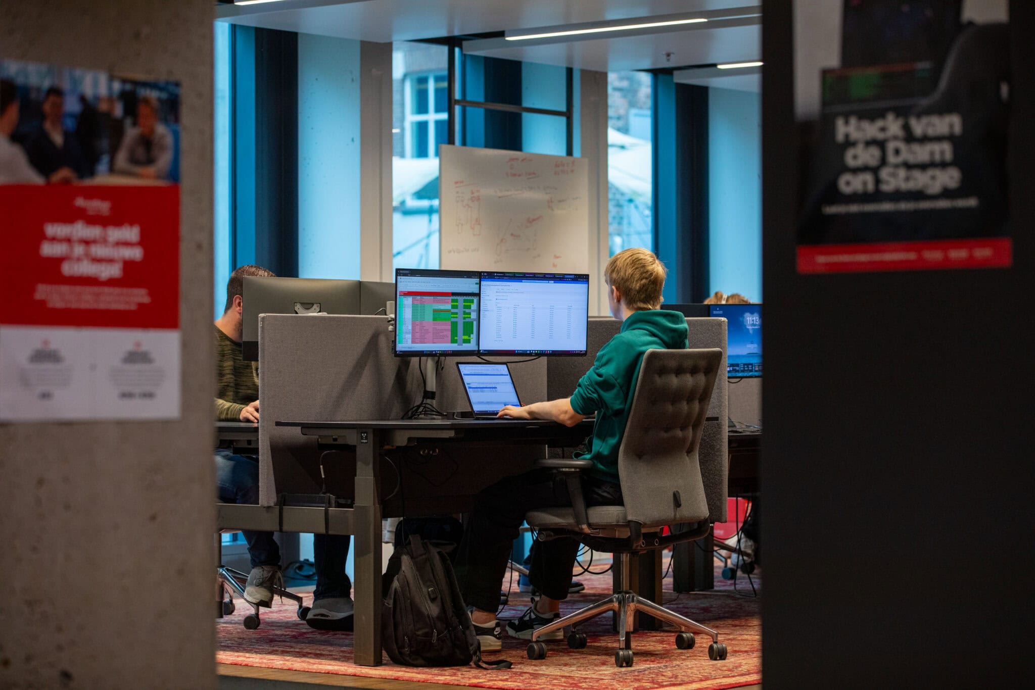 Man op kantoor aan het werk achter een bureau. Te zien zijn twee vervaagde poster op muren en op het bureau staat een laptop met twee extra beeldschermen.